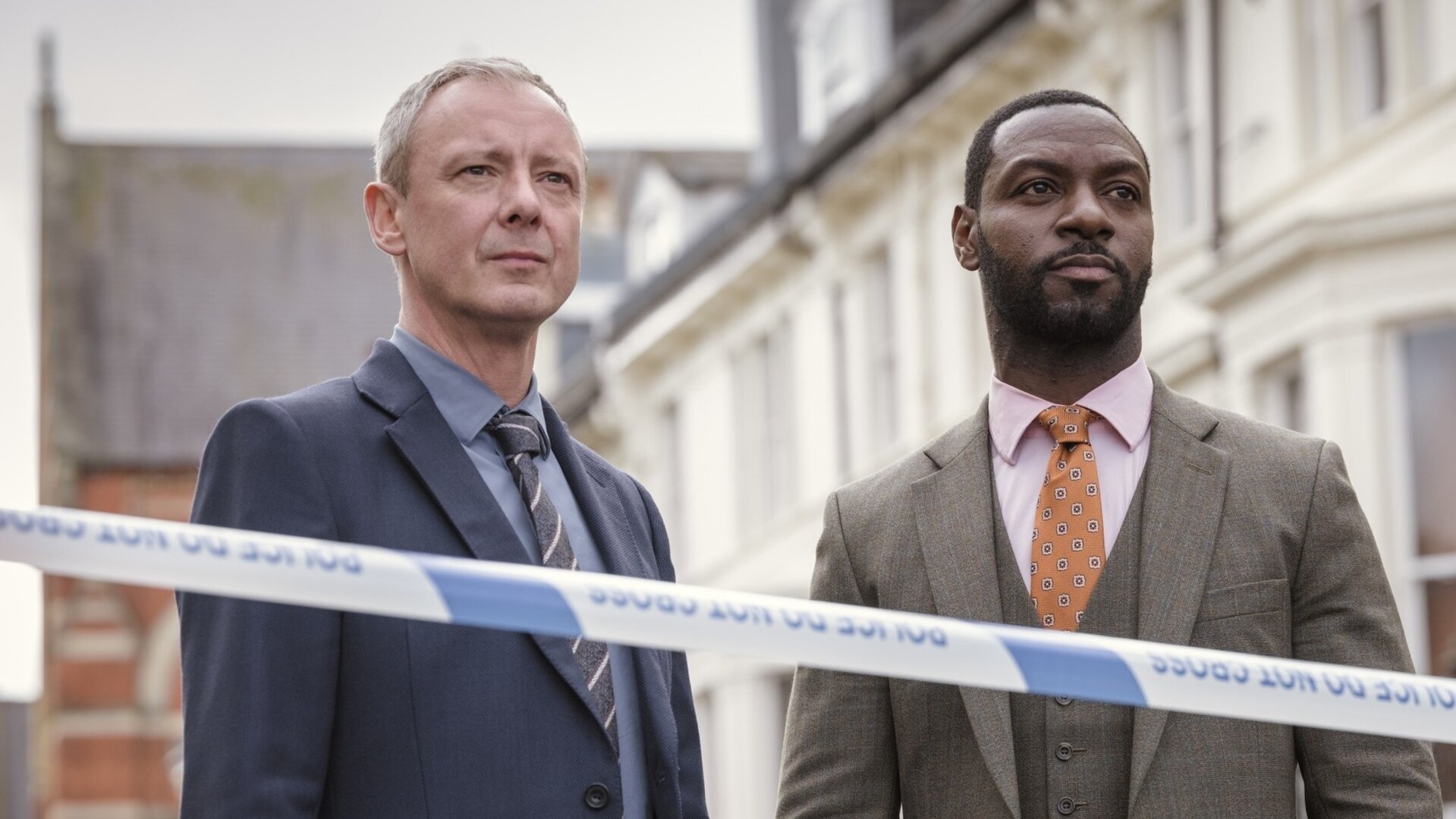 "Detective Grace - Rigor Mortis": DS Roy Grace (John Simm) und DS Glenn Branson (Richie Campbell) stehen nebeneinander auf einer Straße hinter einem Absperrband mit der Aufschrift "Police - do not cross".