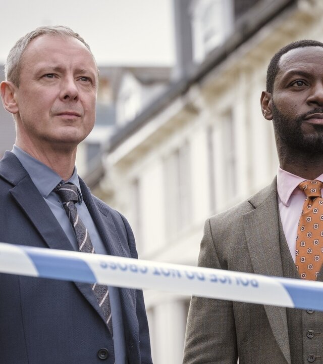 "Detective Grace - Rigor Mortis": DS Roy Grace (John Simm) und DS Glenn Branson (Richie Campbell) stehen nebeneinander auf einer Straße hinter einem Absperrband mit der Aufschrift "Police - do not cross".