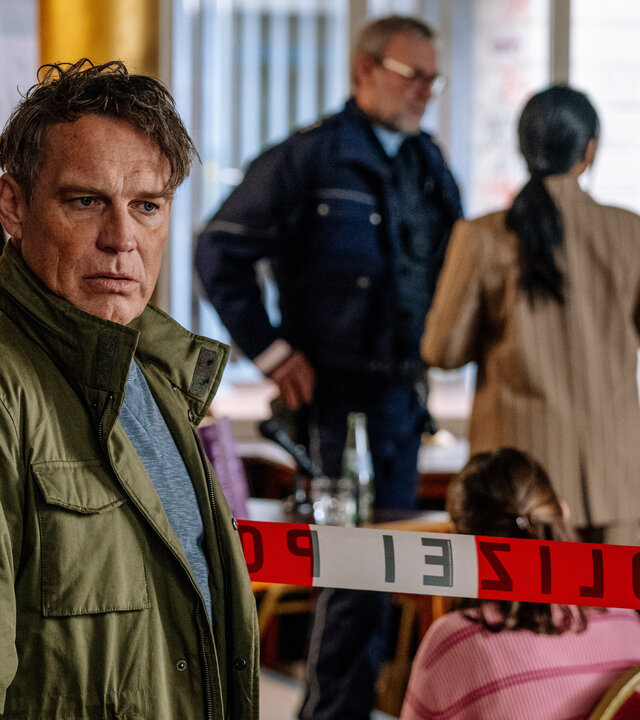 "SOKO KÃ¶ln - TÃ¶dlicher Bestseller": Matti Wagner (Pierre Besson) steht vor einem Polizeiabsperrband in einer Buchhandlung und blickt ernst nach rechts. Hinter dem Absperrband sitzen mehrere junge Personen (Komparsen) auf StÃ¼hlen und ein Streifenpolizist (Komparse) spricht mit einer Frau (Komparsin).