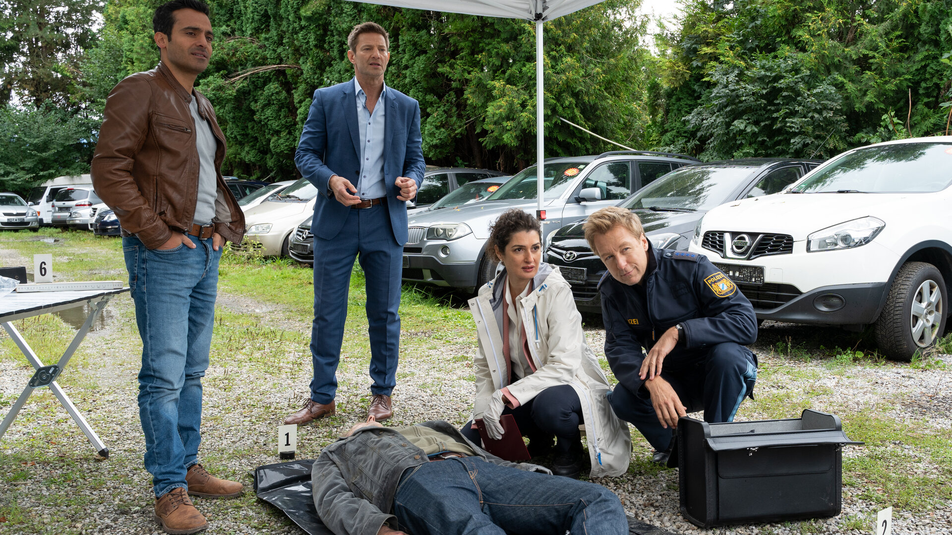 "Die Rosenheim-Cops - Stadlers Stadlfest": Kommissar Kaya (Baran Hêvî), Kommissar Hansen (Igor Jeftić), Dr. Dimos (Anastasia Papadopoulou) und Polizeihauptmeister Mohr (Max Müller) beraten sich am Tatort. Vor ihnen liegt das Opfer (Komparse) rücklings auf einer dunklen Plane. Im Hintergrund stehen diverse Pkw. Vor Polizeihauptmeister Mohr steht der schwarze Arztkoffer von Dr. Dimos.