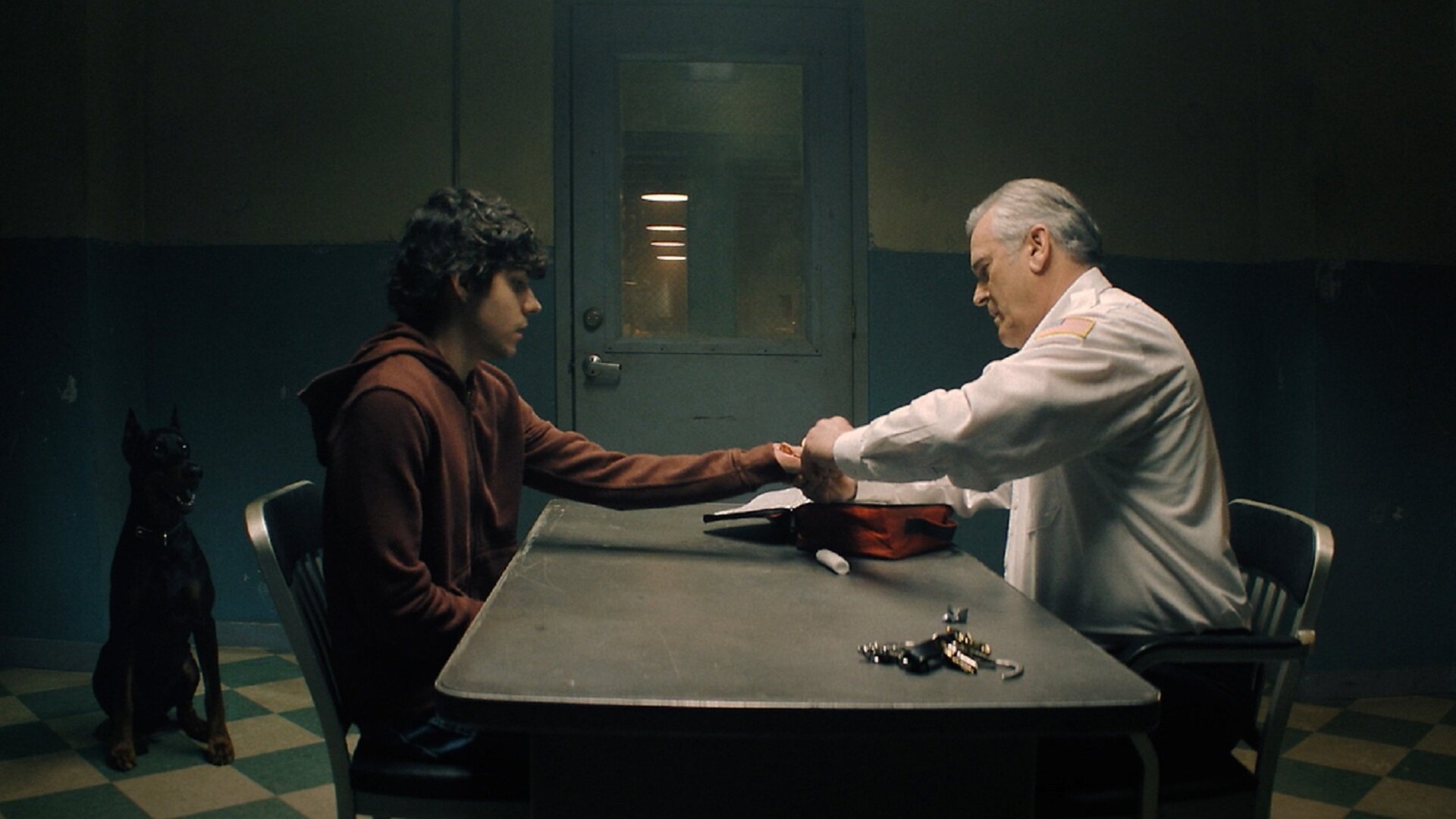 "Hysteria! - Male des Teufels": Chief Dandridge (Bruce Campbell) und Dylan (Emjay Anthony) sitzen sich an einem Tisch in einem karg beleuchteten Raum gegenüber. Dandridge verbindet Dylans Hand. Auf dem Tisch liegt ein Schlüsselbund und eine rote Tasche, neben dem Tisch sitzt ein großer Hund.