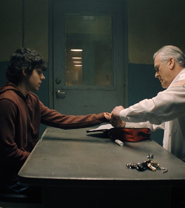 "Hysteria! - Male des Teufels": Chief Dandridge (Bruce Campbell) und Dylan (Emjay Anthony) sitzen sich an einem Tisch in einem karg beleuchteten Raum gegenüber. Dandridge verbindet Dylans Hand. Auf dem Tisch liegt ein Schlüsselbund und eine rote Tasche, neben dem Tisch sitzt ein großer Hund.