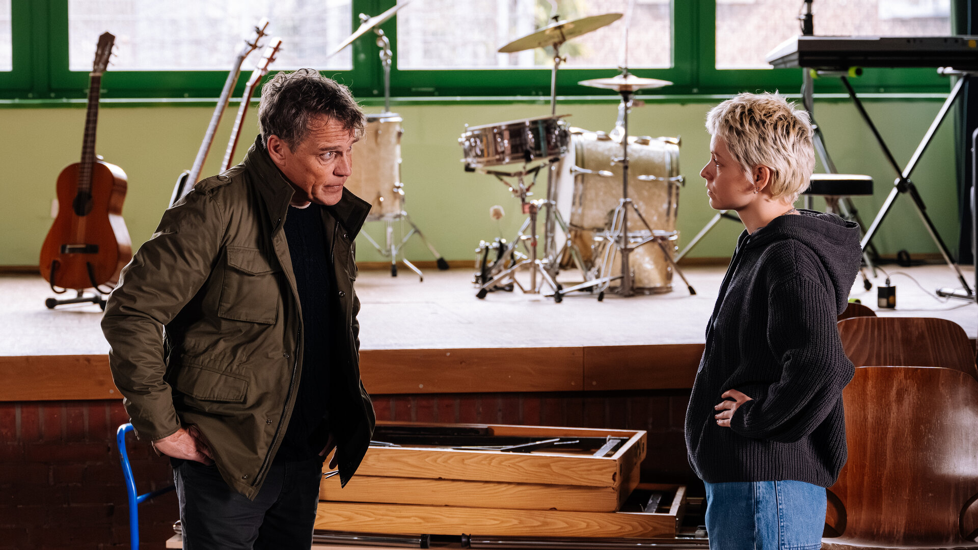 "SOKO KÃ¶ln - Ein Platz zum Sterben": Matti Wagner (Pierre Besson) steht Pia Berbesch (Antonia Breidenbach) vor einer BÃ¼hne, auf der sich diverse Instrumente befinden, gegenÃ¼ber. Er hat die HÃ¤nde in die HÃ¼ften gestemmt und sieht sie auffordernd an. Pia Berbesch spiegelt seine Haltung und erwidert den Blick ausdruckslos. Vor der BÃ¼hne liegen aufeinandergestapelte und zusammengeklappte Holztische auf einem Wagen.