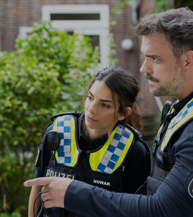 "Notruf Hafenkante - Maria brennt": Die Polizistin Isa Nowak (Lilli Hollunder) und ihr Kollege Kris Freiberg (Marc Barthel) stehen dicht nebeneinander in einem Garten vor einer Marienstatue. Kris weißt mit seinem linken Zeigefinger auf etwas auf der Statue. Die Statue ist verbrannt.