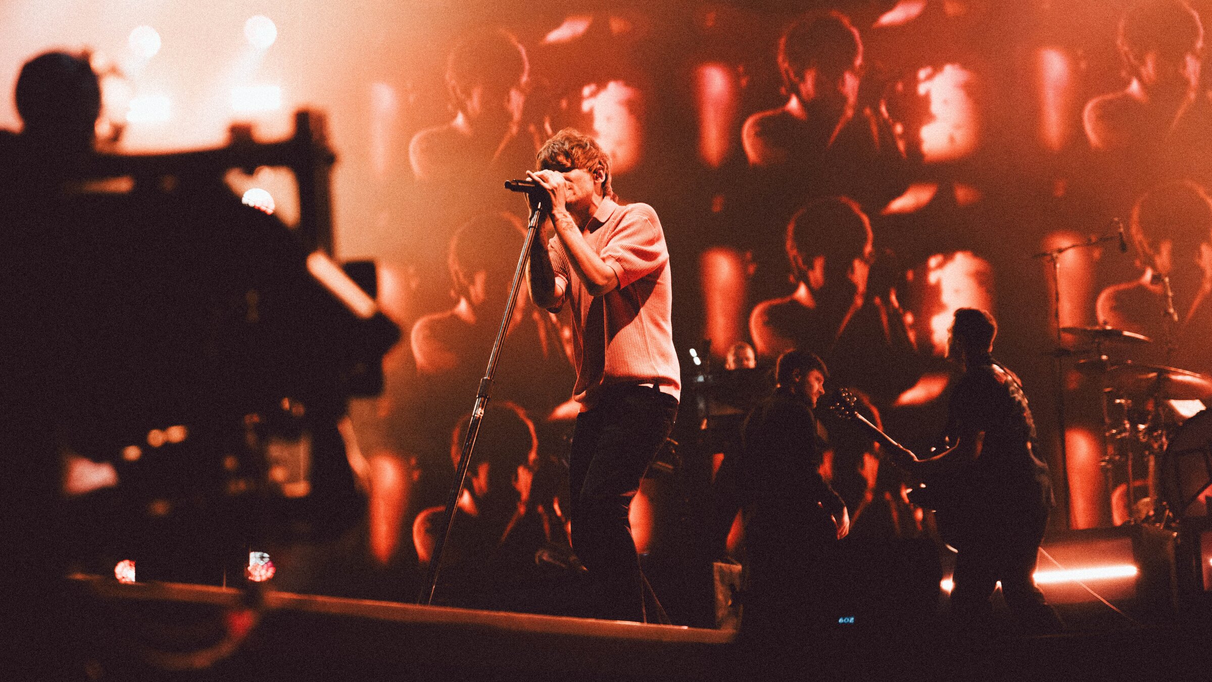 "Louis Tomlinson: For Every Question Why - Live": Der Sänger Louis Tomlinson steht auf der Bühne und singt in ein Mikrofon. Im Hintergrund weitere Musiker der Band. Die Bühnenrückwand wird das Geschehen auf der Bühne projiziert.