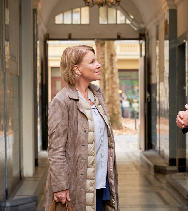 "Marie Brand und der entsorgte Mann": Marie Brand (Mariele Millowitsch) und Jürgen Simmel (Hinnerk Schönemann) stehen zusammen mit Dr. Engler (Thomas Heinze) in einem Gang zu einen Wohnhaus und schauen ihn an. Dr. Engler lächelt die beiden an. Marie Brand hält in ihrer rechten Hand eine beigefarbene Reisetasche.