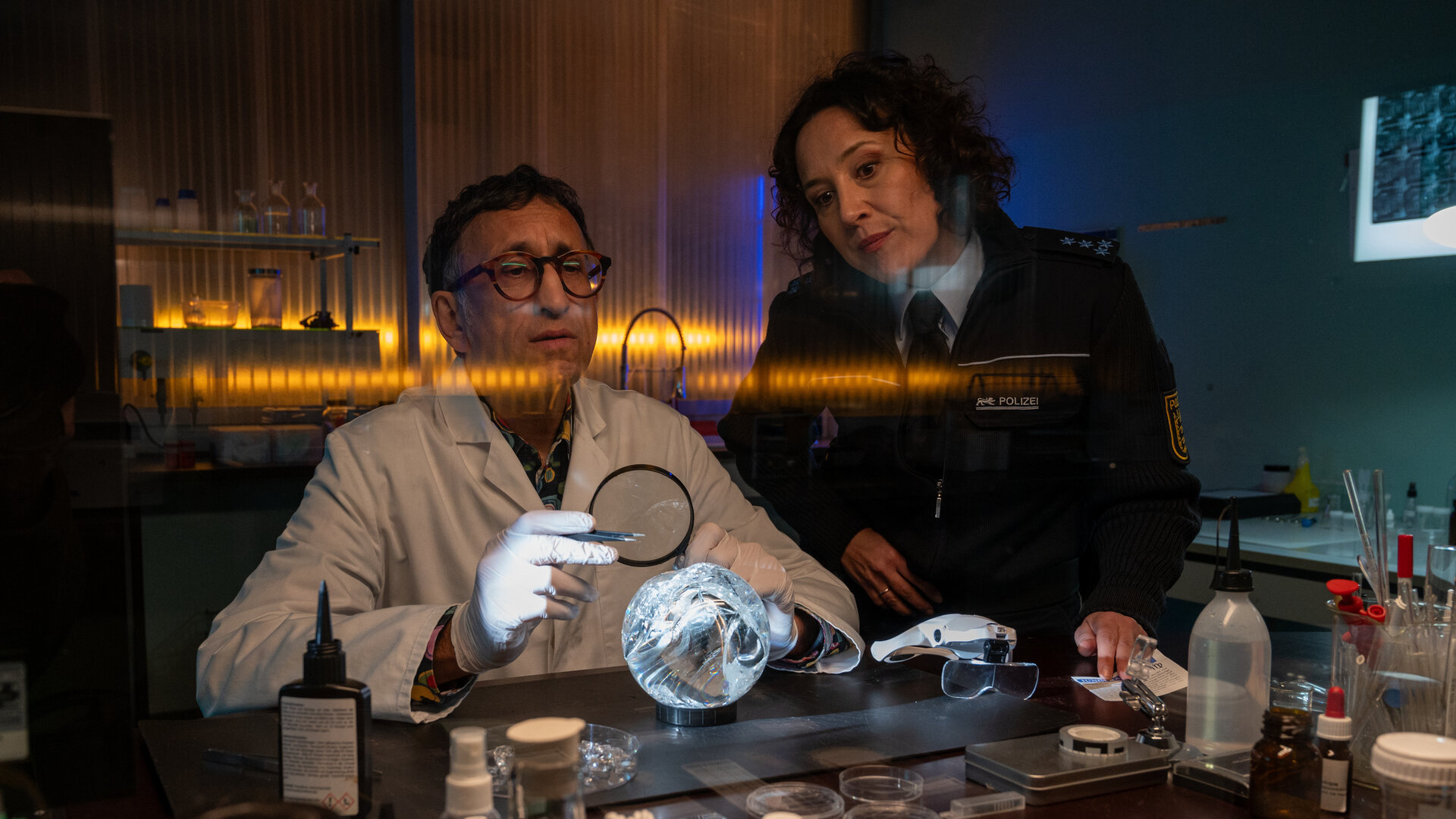 "SOKO Stuttgart - Lauf der Sterne": Der Leiter der KTU Jan Arnaud (Mike Zaka Sommerfeld) sitzt in seinem Labor am Arbeitstisch und untersucht mit einer Lupe und einer Pinzette eine angebrochene Kristallkugel. Neben ihm steht die Streifenbeamtin Bille Beyer (Bärbel Stolz) und schaut ihm kritisch bei der Arbeit zu.