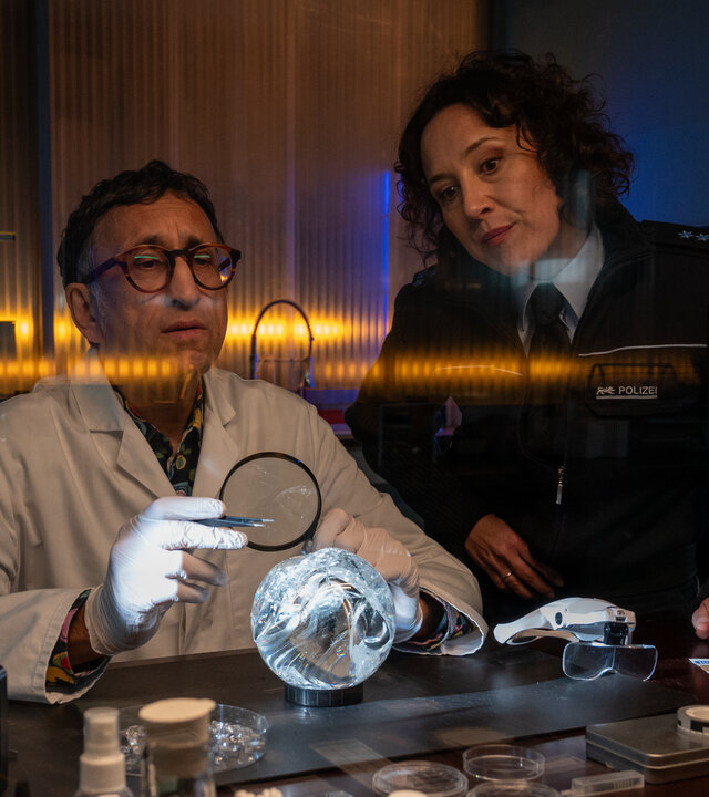 "SOKO Stuttgart - Lauf der Sterne": Der Leiter der KTU Jan Arnaud (Mike Zaka Sommerfeld) sitzt in seinem Labor am Arbeitstisch und untersucht mit einer Lupe und einer Pinzette eine angebrochene Kristallkugel. Neben ihm steht die Streifenbeamtin Bille Beyer (Bärbel Stolz) und schaut ihm kritisch bei der Arbeit zu.