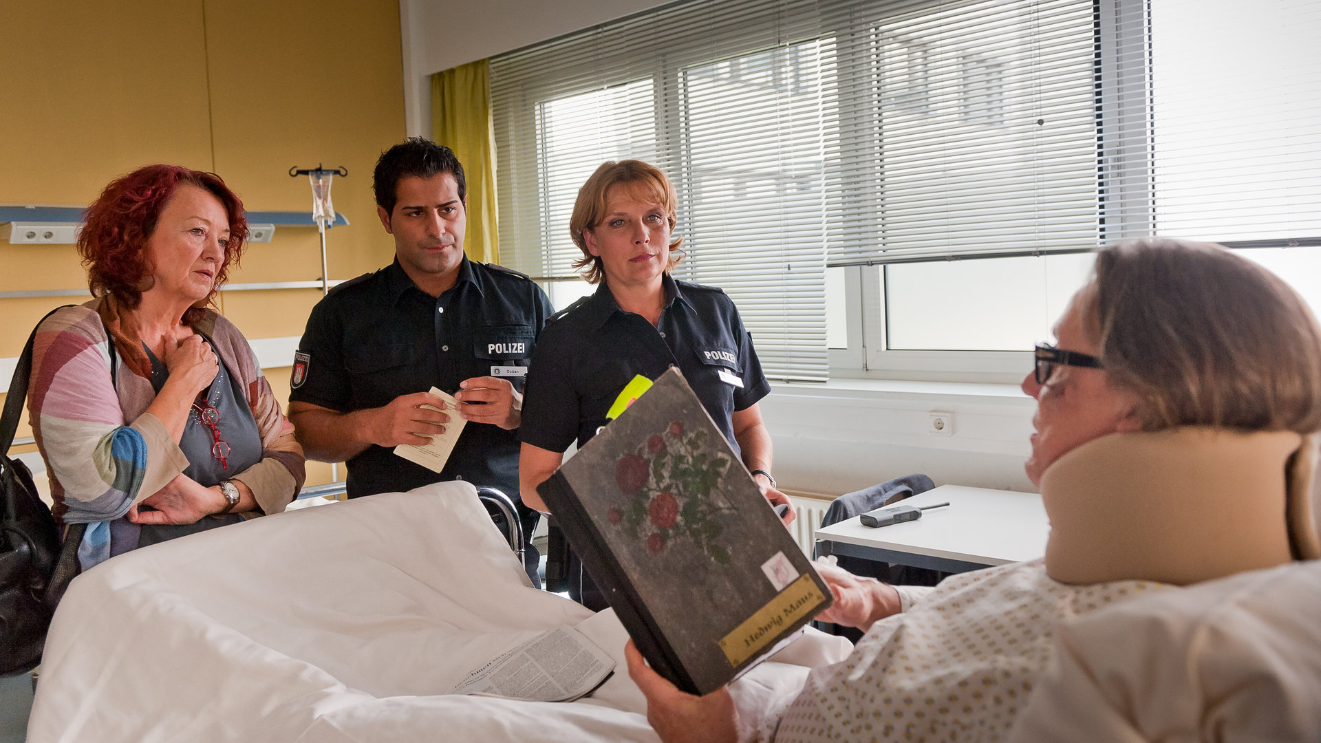 "Notruf Hafenkante - Der letzte Vorhang": Theaterregisseur Harting (Hannes Hellmann) liegt im Krankenhausbett und hält ein Buch in der Hand. Am Fußende des Bettes stehen die Souffleuse Maus (Barbara Focke) sowie die Polizisten Claudia (Janette Rauch) und Tarik (Serhat Cokgezen).