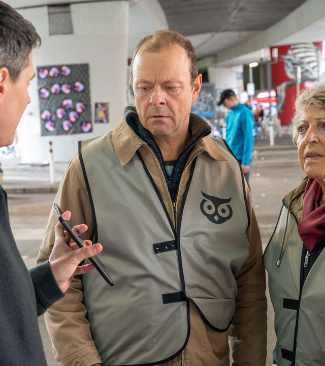"SOKO Stuttgart - Die Nachtwache": Unter einer Stuttgarter Stadtbrücke befragt Kriminalkommissar Rico Sander (Benjamin Strecker) Hanni Walter (Marie-Luise Marjan) und Volker Schwelk (Hanns-Jörg Krumpholz). Er hält ihnen sein Handy entgegen. Die beiden tragen über ihrer Privatkleidung graue Westen, auf denen ein Eulenkopf, das Logo der "Nachtwache", aufgedruckt ist. Im Hintergrund sind weitere Mitglieder der "Nachtwache" dabei, Müll aufzusammeln.