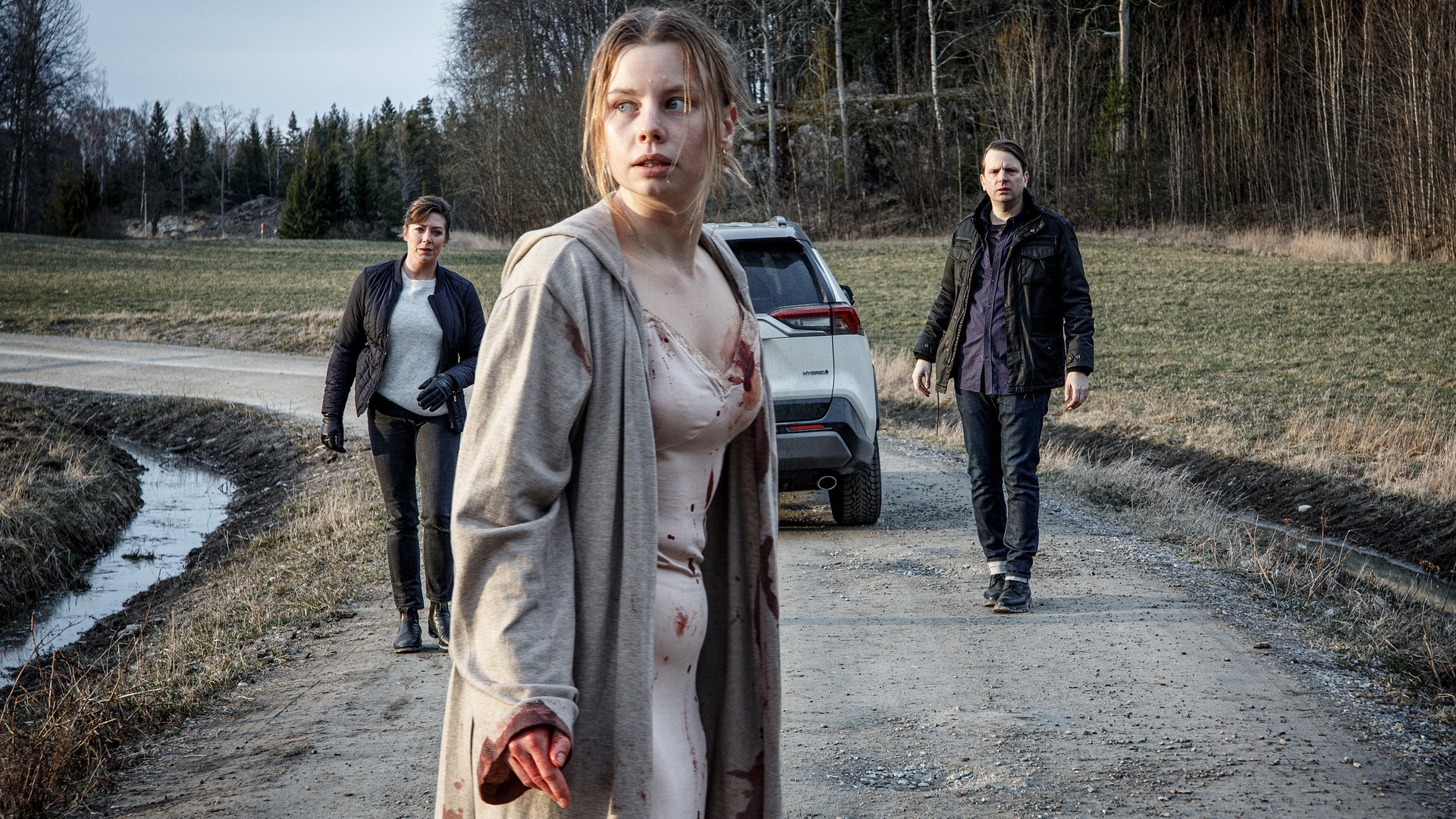 "Kommissar Beck - Ohne jeden Zweifel": Leichtbekleidet und blutverschmiert steht Ida Lind (Amanda Jansson) mitten auf einem Feldweg und blickt panisch um sich. Alexandra Beijer (Jennie Silfverhjelm) und Oskar Bergman (Måns Nathanaelsson) sind aus dem Auto ausgestiegen und nähern sich ihr vorsichtig.