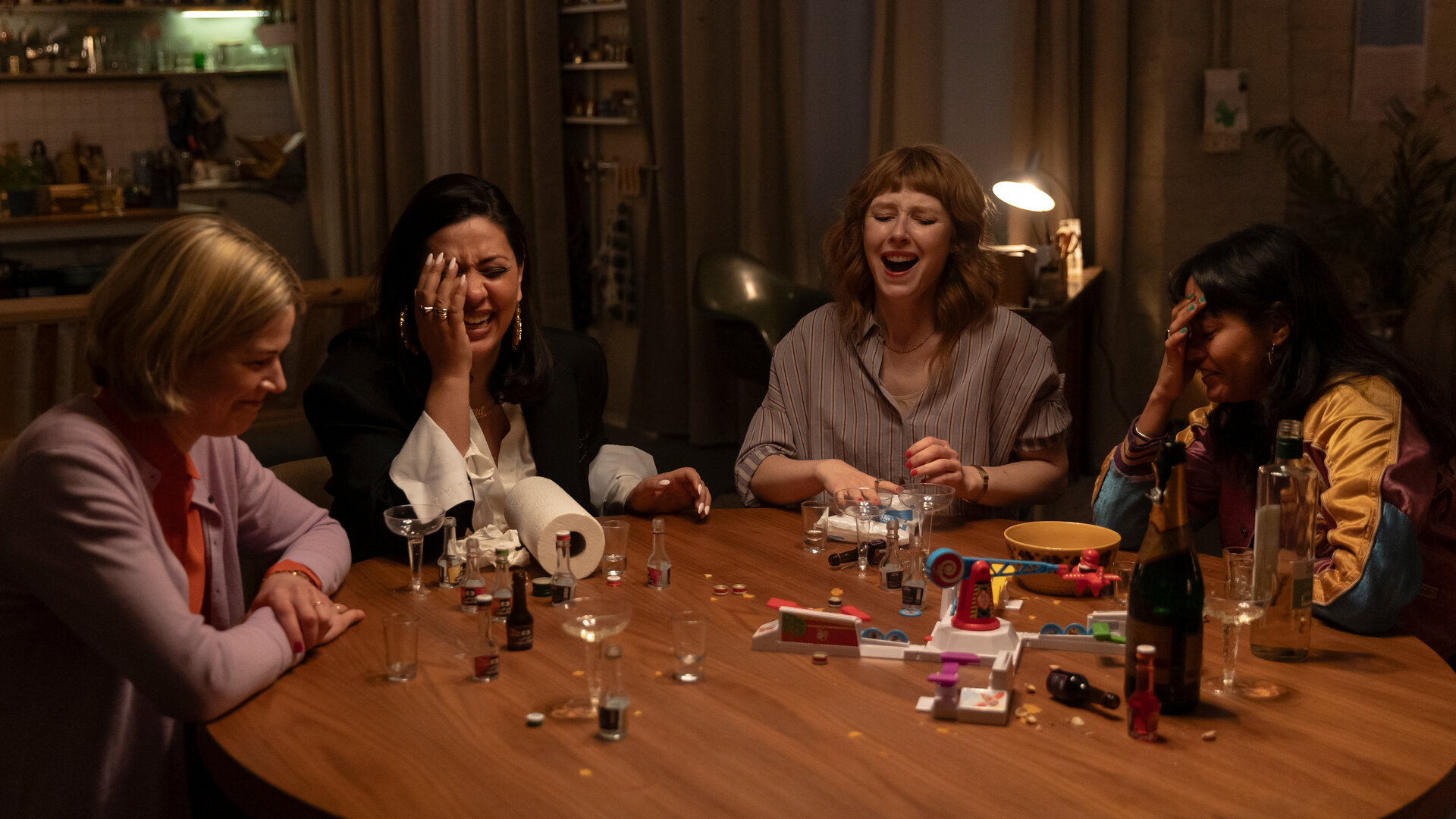 "Deadlines - Die Elektrische": Franzi (Llewellyn Reichman), Elif (Jasmin Shakeri), Lena (Sarah Bauerett) und Jo (Salka Weber) sitzen lachend an einem runden Tisch, auf dem kleine Schnapsflaschen, Gläser und eine Flasche Sekt stehen.