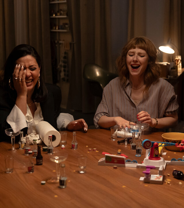 "Deadlines - Die Elektrische": Franzi (Llewellyn Reichman), Elif (Jasmin Shakeri), Lena (Sarah Bauerett) und Jo (Salka Weber) sitzen lachend an einem runden Tisch, auf dem kleine Schnapsflaschen, Gläser und eine Flasche Sekt stehen.