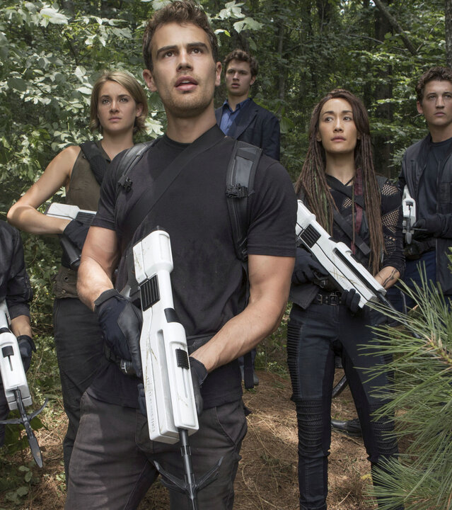 "Die Bestimmung – Allegiant": Four (Theo James) steht zusammen mit seinen Verbündeten, Tris (Shailene Woodley), Peter (Miles Teller), Caleb (Ansel Elgort), Tori (Maggie Q) und Christina (Zoë Kravitz), im Wald. Alle tragen sportliche, dunkle Kleidung und halten futuristische Waffen in den Händen. Verwundert blicken sie in dieselbe Richtung.