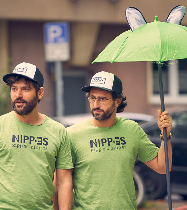 "Andere Eltern 2 – Episode 6": Jannos (Jasin Challah) und Björn (Serkan Kaya) stehen in gebrandeten T-Shirts von "Nipp-es" an ihrem Smoothie-Stand.