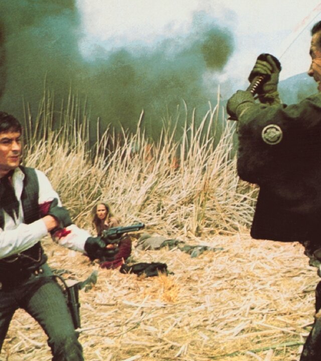 "Rivalen unter roter Sonne": Gauche (Alain Delon) steht mit blutverschmiertem Arm und gezogener Pistole in einem Feld aus trockenem Gras, während Kuroda (Toshirô Mifune) ihm mit erhobenem Schwert gegenübersteht. Im Hintergrund sitzt Christina (Ursula Andress) geduckt zwischen den Gräsern, während dunkler Rauch aufsteigt.