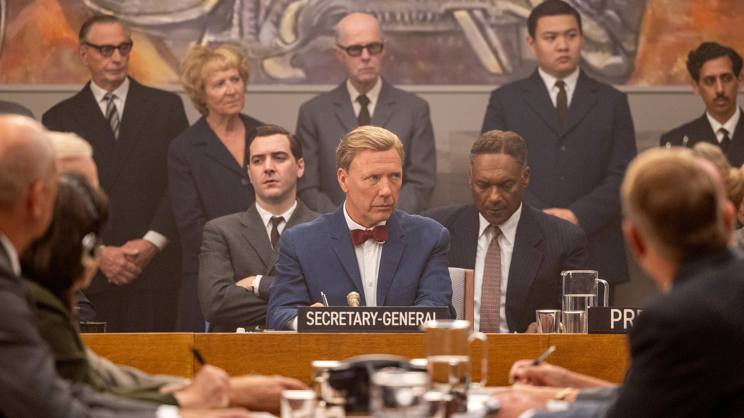„Hammarskjöld – Kampf für den Frieden“: In der Bildmitte sieht man Dag Hammarskjöld (Mikael Persbrandt) während einer Versammlung der UN. Vor ihm ist ein Schild mit seiner Amtsbezeichnung „Secretary-General“. Direkt seitlich von ihm sitzen seine Berater Heinrich Wieschoff (Cian Barry, links) und Ralph Bunche (Colin Salmon). Um sie herum befinden sich noch andere Teilnehmer der Versammlung.