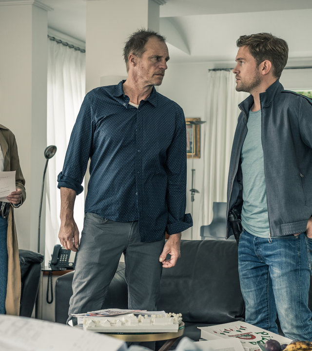 "SOKO München - Notruf": Der rachsüchtige Matthias Berger (Thorsten Nindel) steht in seinem etwas unordentlichen Wohnzimmer. Er beugt sich herausfordernd zu Dominik Morgenstern (Joscha Kiefer), der neben ihm steht und beide Hände in den hinteren Hosentaschen hat. Beide Männer sehen sich ernst in die Augen. Resi Schwaiger (Mersiha Husagic) steht einen Schritt hinter Matthias Berger. Während sie ihn beobachtet, hält sie einen aufgefalteten Brief in den Händen.