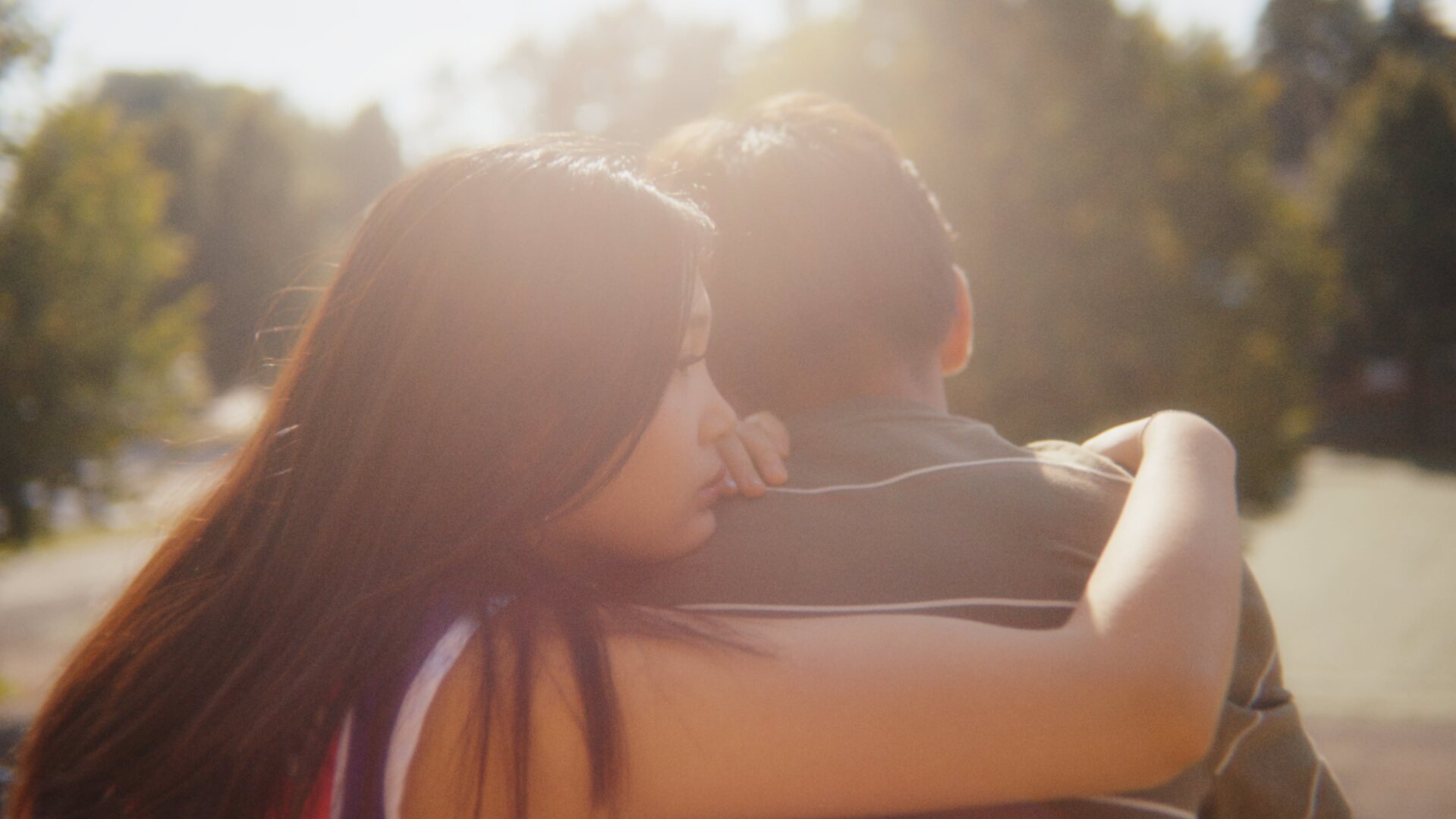 "After Benjamin: Das Konzert": Lina (Victoria Hoang) sitzt neben Elias (Sam Ashraf) im Freien mit dem Rücken zur Kamera auf einer Bank. Das Wetter ist schön, die Sonne scheint von vorn auf die beiden. Lina sitzt Elias zugewandt, umarmt ihn mit ihrem rechten Arm und lehnt ihr Kinn auf seine Schulter. Elias schaut geradeaus, den Kopf leicht nach unten gebeugt.