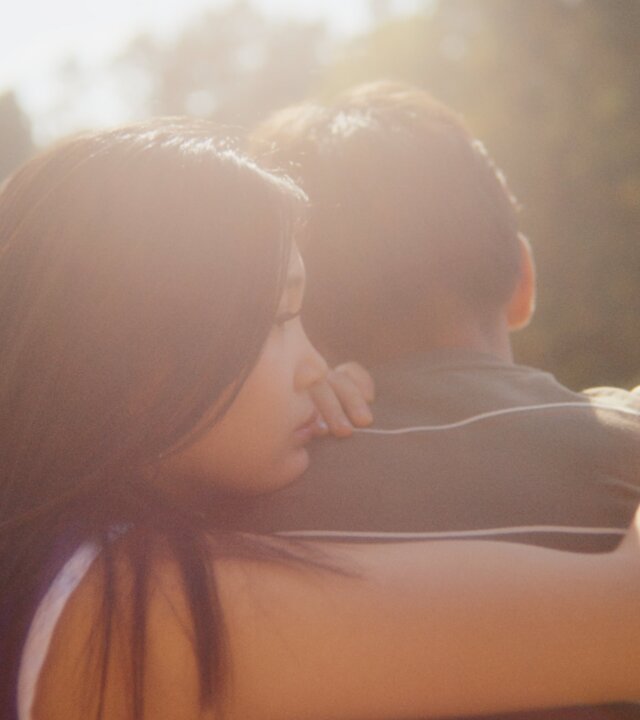 "After Benjamin: Das Konzert": Lina (Victoria Hoang) sitzt neben Elias (Sam Ashraf) im Freien mit dem Rücken zur Kamera auf einer Bank. Das Wetter ist schön, die Sonne scheint von vorn auf die beiden. Lina sitzt Elias zugewandt, umarmt ihn mit ihrem rechten Arm und lehnt ihr Kinn auf seine Schulter. Elias schaut geradeaus, den Kopf leicht nach unten gebeugt.