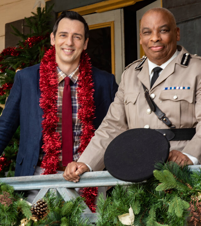 "Death in Paradise - Der Weihnachtsgeist (1)": Detective Neville Parker (Ralf Little) und der Polizeichef Selwyn Patterson (Don Warrington) stehen auf der weihnachtlich dekorierten Veranda der Polizeiwache und schauen zufrieden in die Ferne.