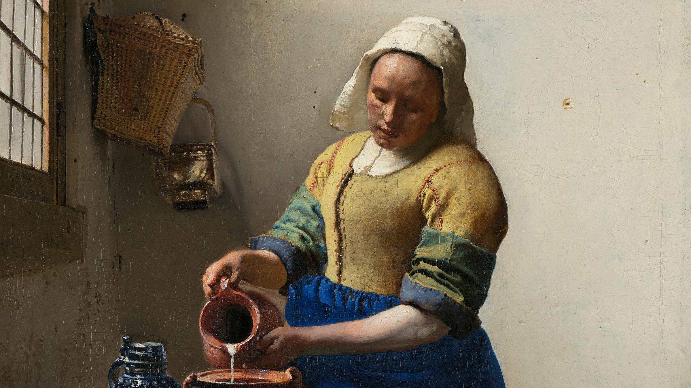 "Kunst für die Ewigkeit: Jan Vermeer": Eine Frau in historischer Kleidung gießt Milch aus einem Krug in eine Schale auf einem mit Brot und Keramikgefäßen gedeckten Tisch vor einem Fenster.