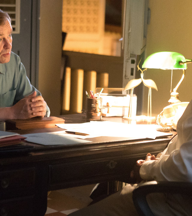 "Death in Paradise - Der letzte Trumpf": Commissioner Selwyn Patterson (Don Warrington) und DI Jack Mooney (Ardal O''Hanlon) sitzen sich am Schreibtisch gegenüber und führen ein Gespräch.