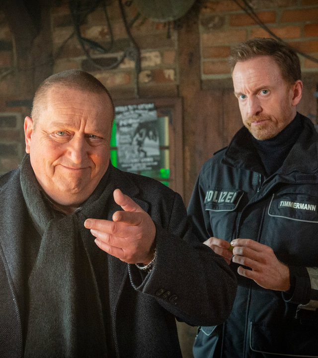 "SOKO Wismar - Ilse muss weg": Ein Spielautomat hängt an der Backsteinwand der Kneipe. Reuter (Udo Kroschwald) macht ein verlegenes Gesicht während er mit dem Finger auf sein Gegenüber deutet, das dem Betrachter verborgen bleibt. Timmermann (Mathias Junge) steht hinter Reuter und blickt entschuldigend drein.