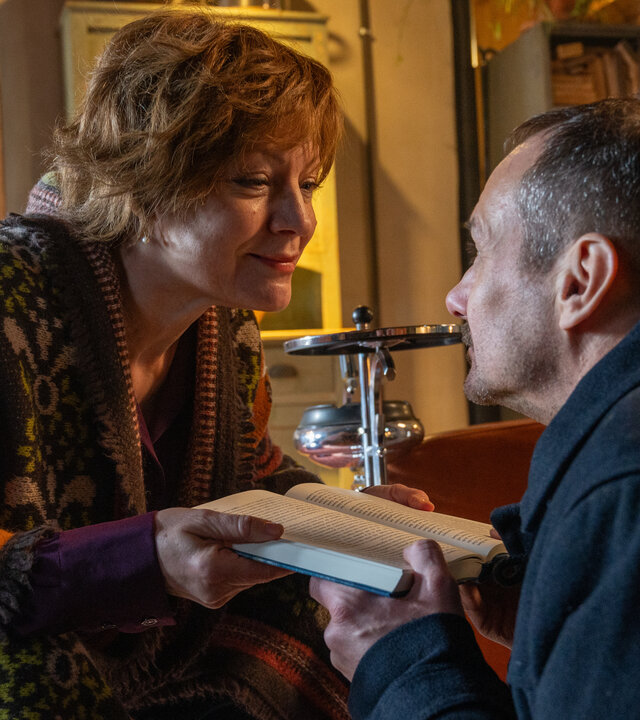 "SOKO Stuttgart - Druck": Die Autorin Charlotte Ghetta (Marion Mitterhammer) und ihr Agent Fabian Steffen (Knut Berger) hocken sich in einem Wohnraum gegenüber. Beide halten ein aufgeschlagenes Buch zwischen sich fest und schauen sich lächelnd tief in die Augen.