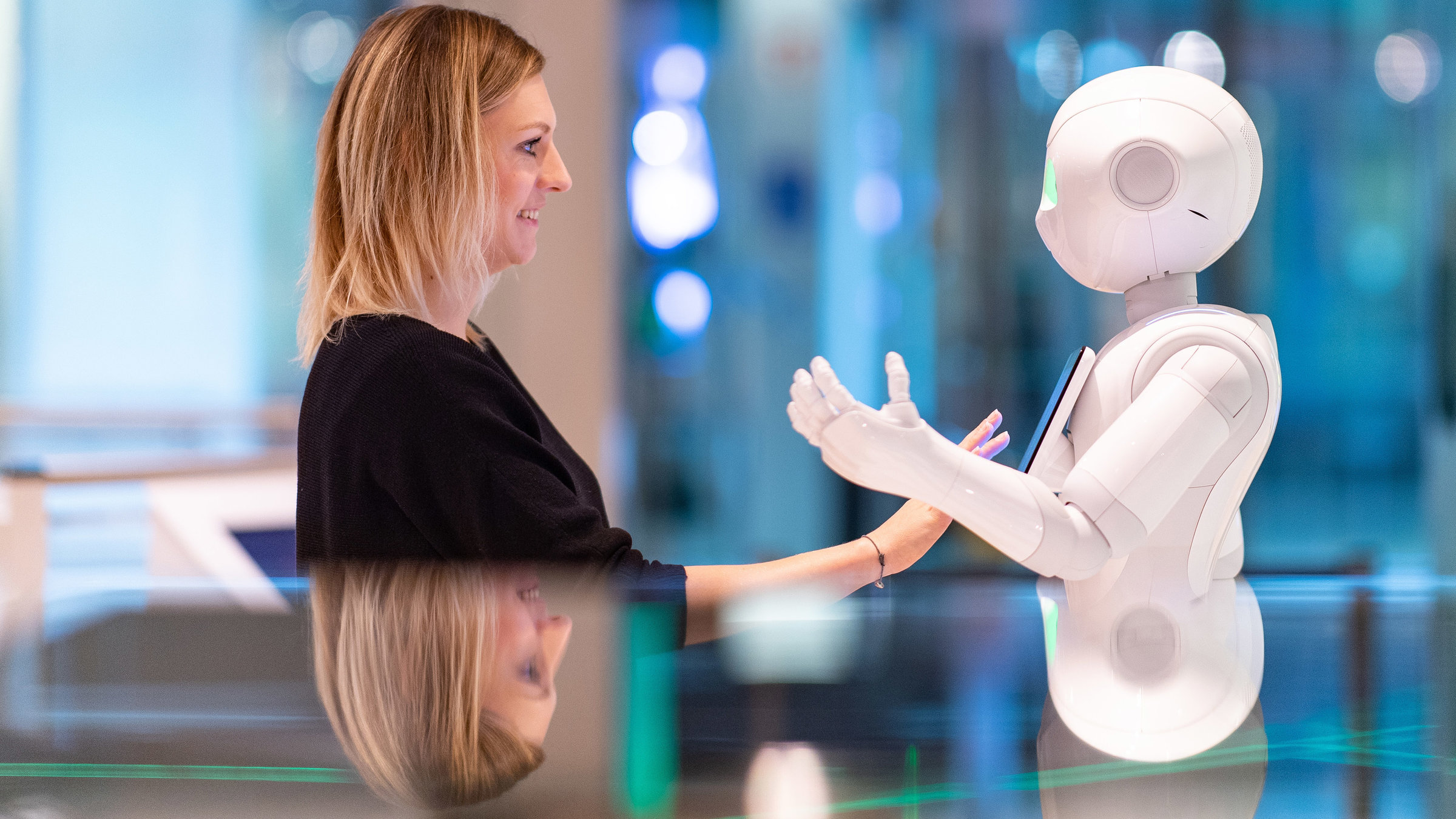 "sonntags - Deutschland verändert sich: Zukunftsvisionen": Eine Frau steht einem Service-Roboter in Menschengestalt gegenüber und hält die Hand lächelnd vor ein Display, das sich auf seiner Brust befindet. Der Roboter hat die linke Hand erhoben.