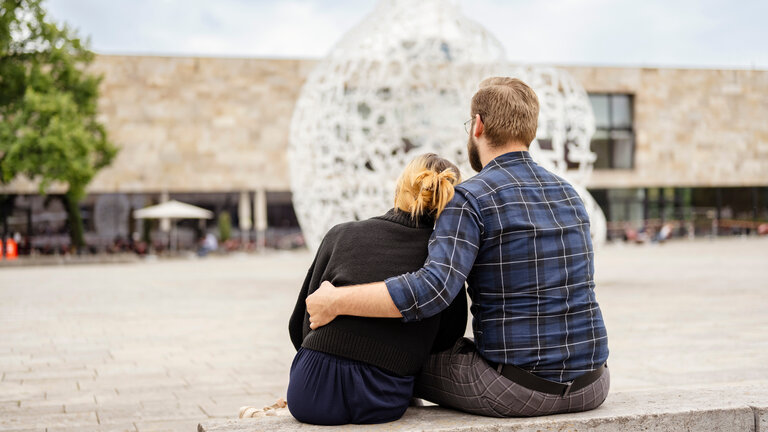 "37°Leben: Arm an der Uni - Wenn das Studium alles kostet": Selina und Dennis sitzen mit dem Rücken zur Kamera auf einer Bank auf einem Platz.  Sie sitzen Arm in Arm nebeneinander.