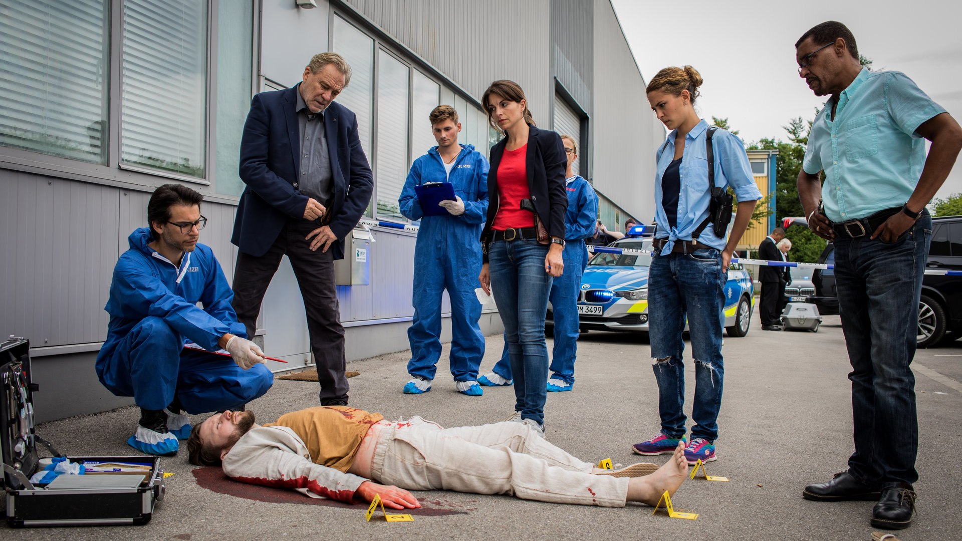 "SOKO München - 24 Stunden": Am Tatort auf einem grauen Industriegelände liegt eine Leiche auf dem Boden. Um diese herum steht das SOKO-Team bestehend aus Arthur Bauer (Gerd Silberbauer), Katharina Hahn (Bianca Hein), Toni Bischoff (Amanda da Gloria) und Franz Ainfachnur (Christofer v. Beau) und schauen sich die Leiche näher an. Dr. Weissenböck (Florian Odendahl) sitzt in der Hocke am Kopf der Leiche, er hält einen Stift in der Hand. Im Hintergrund sind zwei Mitarbeiter der Spusi (Komparsen) und ein Polizeiauto zu sehen.
