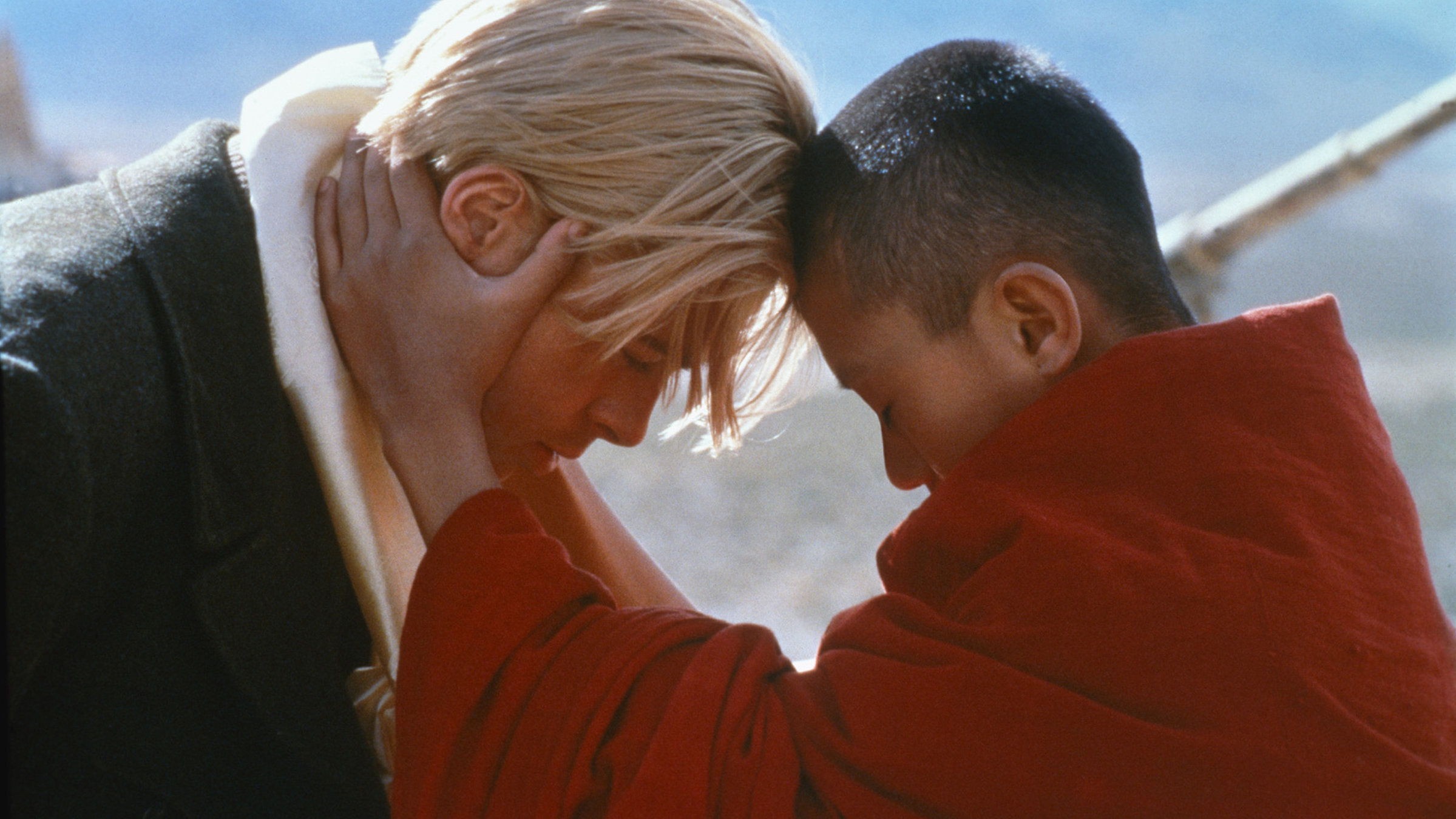 "Sieben Jahre in Tibet": Heinrich Harrer (Brad Pitt), ein junger Mann mit weißblondem Haar, grauem Mantel und weißem Schal, steht dem jugendlichen in ein rotes Gewand gekleideten Dalai Lama gegenüber. Der Jugendliche Tibeter mit kurz geschorenem Haar hält Harrers Kopf in seinen Händen und zieht dessen Stirn zu seiner. Im Hintergrund erkennt man verschwommen ein auf die tibetische Landschaft gerichtetes Fernrohr.