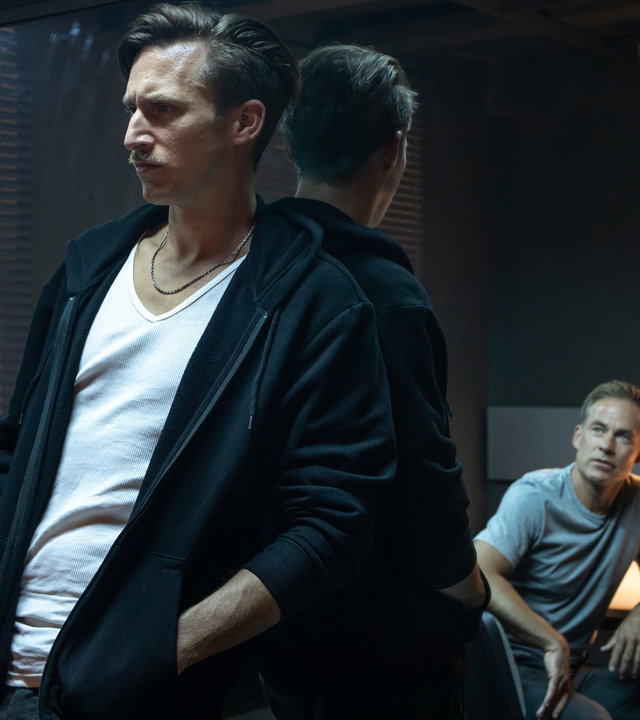 "SOKO Leipzig - Das ewige Leben (2)": Moritz Brenner (Johannes Hendrik Langer) lehnt an einer verspiegelten Scheibe im Vernehmungsraum. Jan Maybach (Marco Girnth) sitzt ihm gegenüber, sodass er nur als Spiegelbild zu erkennen ist.