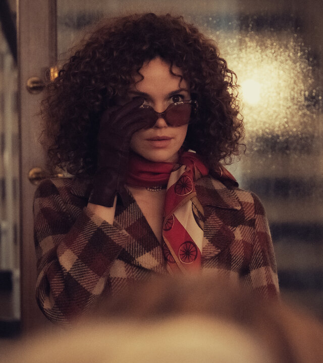 "Ku'damm 77": Im Türrahmen des Saals steht Eva Fassbender (Emilia Schüle). Sie trägt eine Sonnenbrille und zieht diese leicht von den Augen.