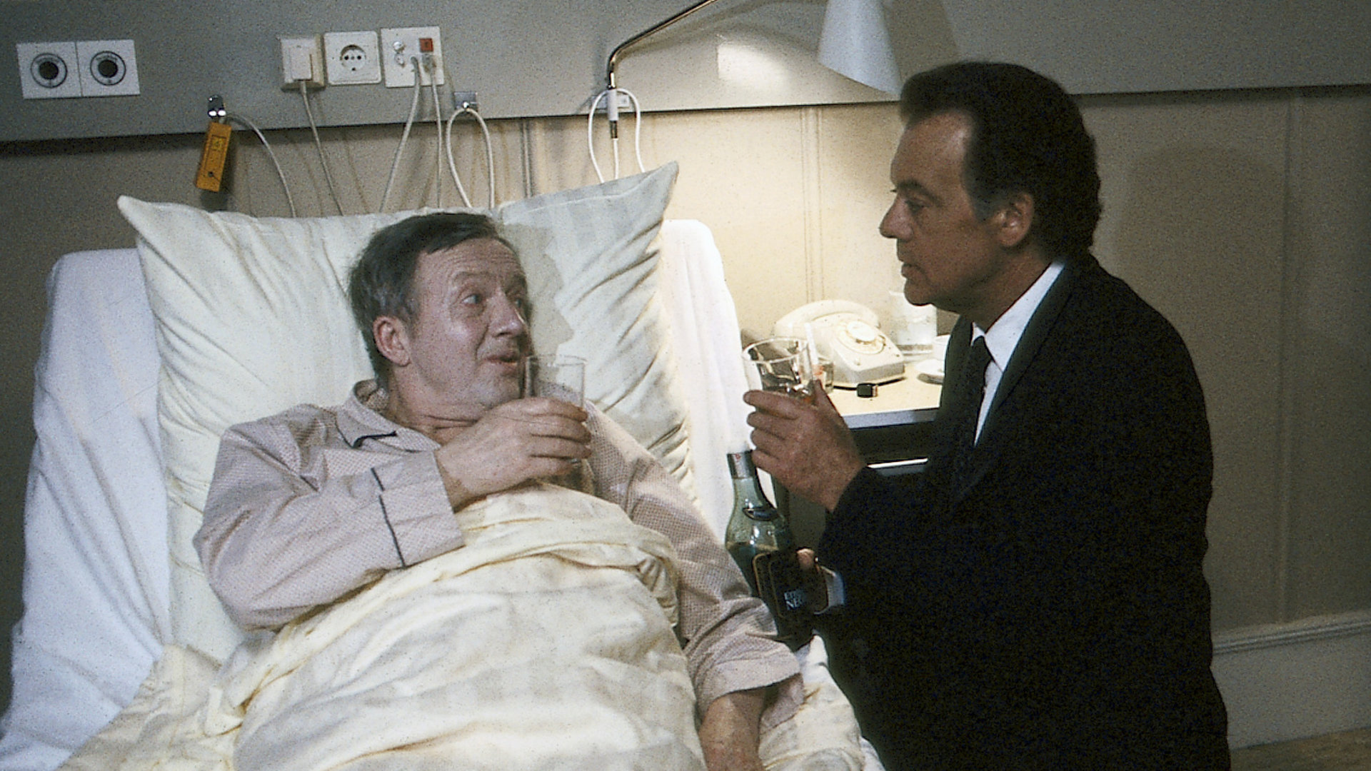 "Die Schwarzwaldklinik - Prost, Herr Professor!": Josef Lambert (Hans-Jürgen Diedrich) liegt im Krankenbett. Neben ihm Prof. Brinkmann (Klausjürgen Wussow). Sie stoßen mit einem Glas Sekt an.