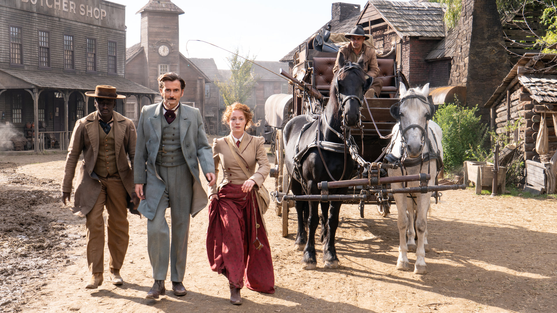 "In 80 Tagen um die Welt (7)": Phileas Fogg (David Tennant), Abigail Fix (Leonie Benesch) und Passepartout (Ibrahim Koma) laufen entschlossen über eine staubige Straße in Battle Mountain, rechts hinter ihnen steht die Pferdekutsche, mit der sie dort eingetroffen sind.