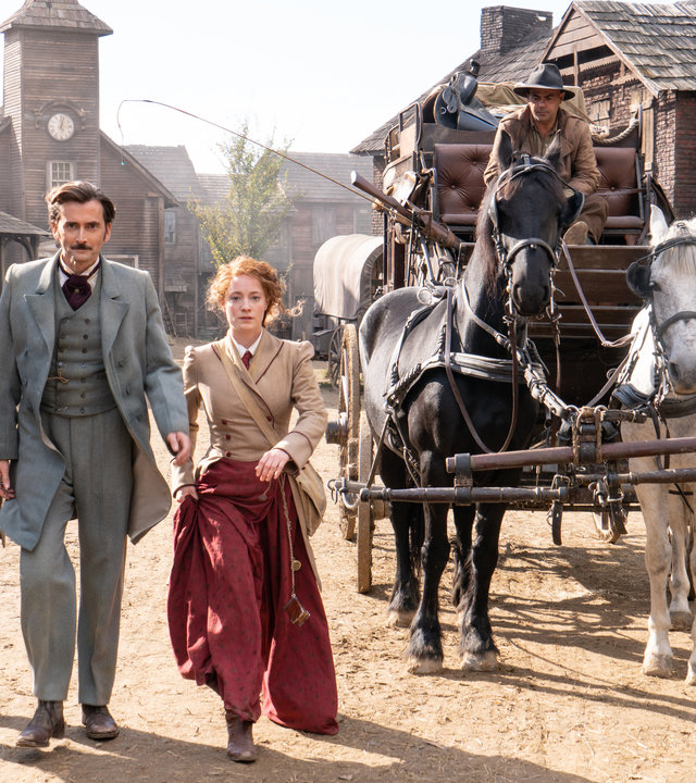 "In 80 Tagen um die Welt (7)": Phileas Fogg (David Tennant), Abigail Fix (Leonie Benesch) und Passepartout (Ibrahim Koma) laufen entschlossen über eine staubige Straße in Battle Mountain, rechts hinter ihnen steht die Pferdekutsche, mit der sie dort eingetroffen sind.