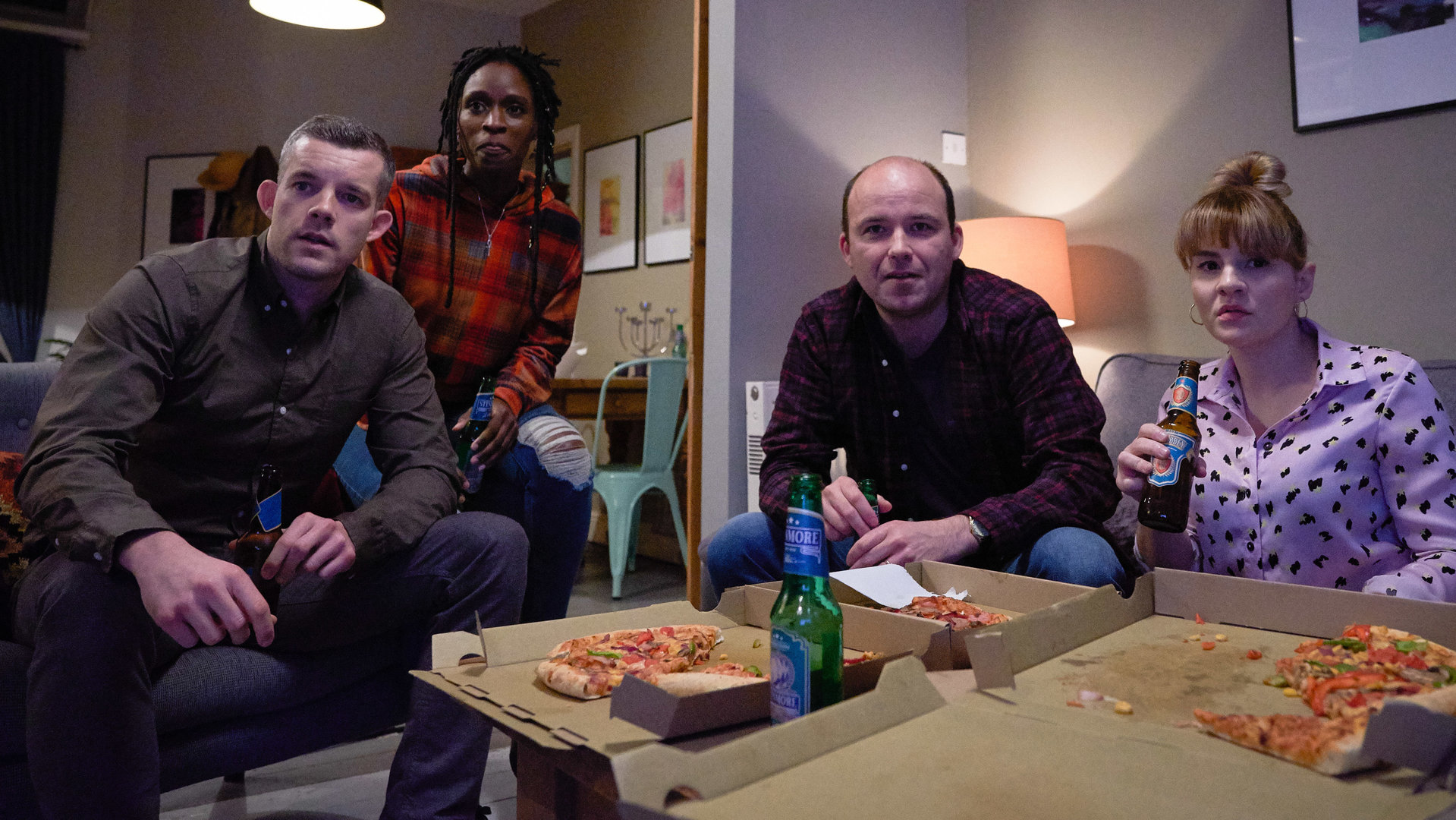 "Years and Years": Daniel Lyons (Russell Tovey), Fran Baxter (Sharon Duncan Brewster), Stephen Lyons (Rory Kinnear) und Rosie Lyons (Ruth Madeley) sitzen zusammen im Wohnzimmer und essen Pizza.