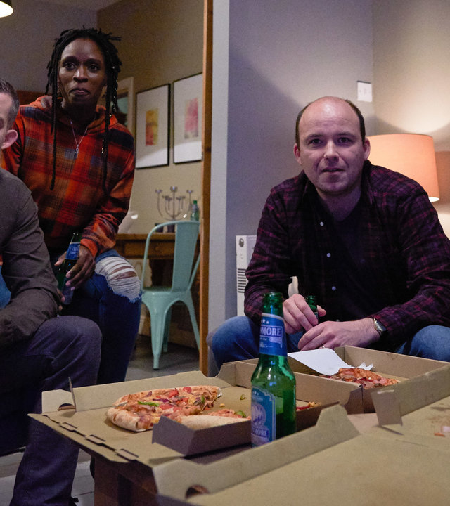 "Years and Years": Daniel Lyons (Russell Tovey), Fran Baxter (Sharon Duncan Brewster), Stephen Lyons (Rory Kinnear) und Rosie Lyons (Ruth Madeley) sitzen zusammen im Wohnzimmer und essen Pizza.
