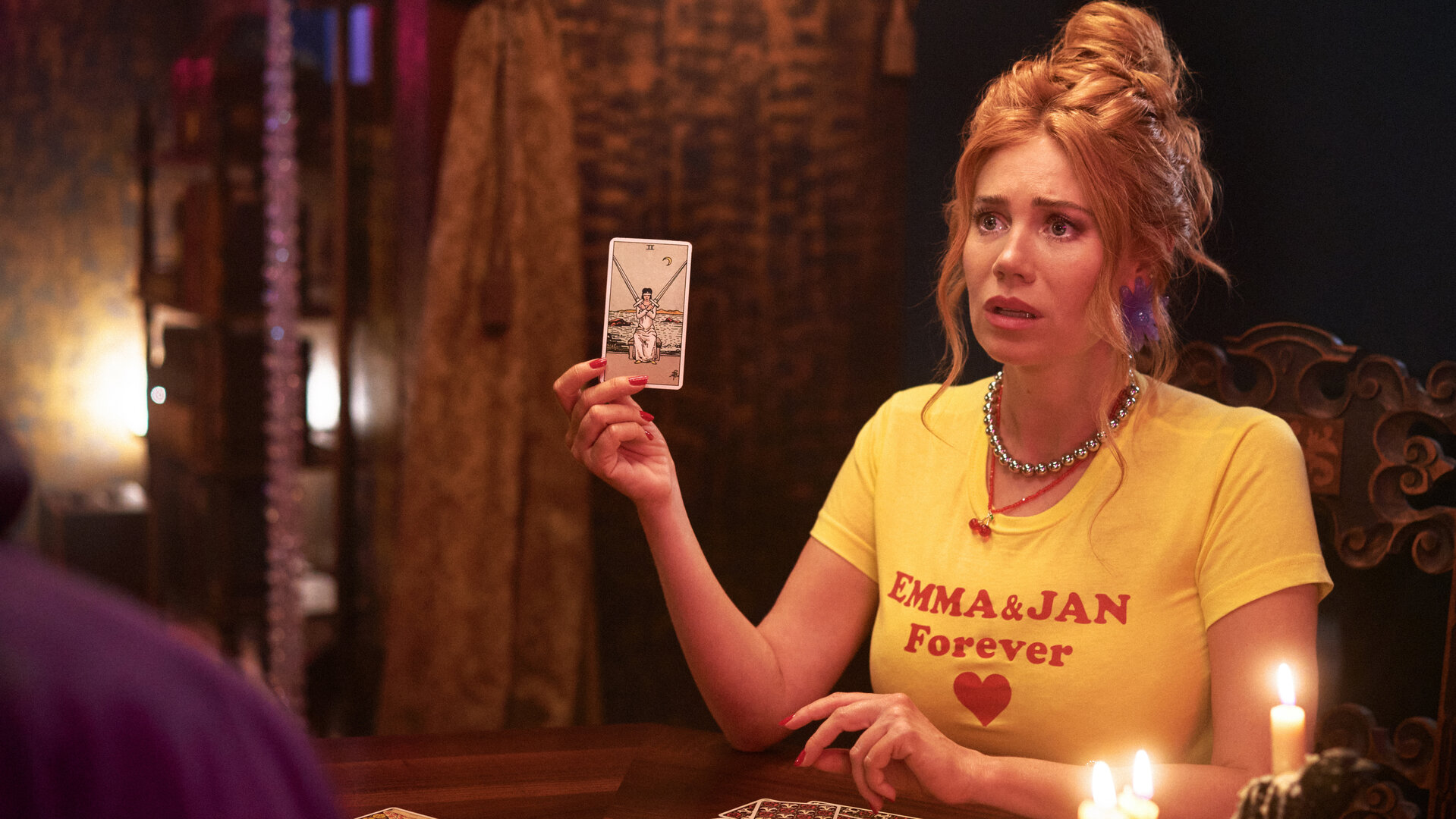 "My Ex - Paul": Ivy (Palina Rojinski) sitzt in einem dunklen Raum am Tisch und hat aus all den Karten, die auf dem Tisch liegen, eine herausgezogen. Sie hält sie mit ihrer rechten Hand nach oben. Auf dem Tisch stehen brennende Kerzen, im Hintergrund spendet eine Wandleuchte spärliches Licht.