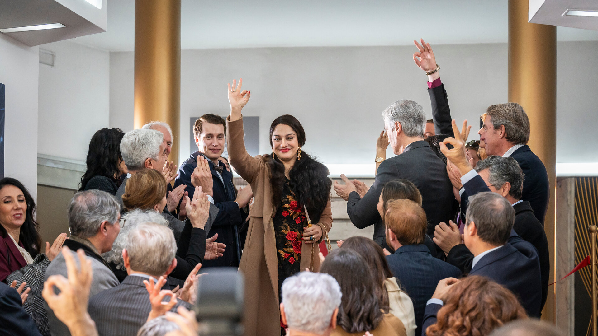 "Take the Money and Run - One Coin": Ruja Ignatova (Nilam Farooq) und Jonas Kurth (László Branko Breiding) stehen gemeinsam mit vielen anderen Personen auf einer Eingangstreppe. Ruja und einige andere Personen bilden mit Daumen und Zeigefinder einen Kreis. Die Stimmung ist sehr lebhaft.