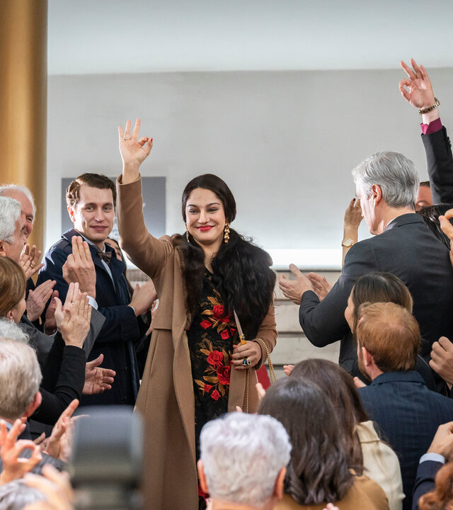 "Take the Money and Run - One Coin": Ruja Ignatova (Nilam Farooq) und Jonas Kurth (László Branko Breiding) stehen gemeinsam mit vielen anderen Personen auf einer Eingangstreppe. Ruja und einige andere Personen bilden mit Daumen und Zeigefinder einen Kreis. Die Stimmung ist sehr lebhaft.