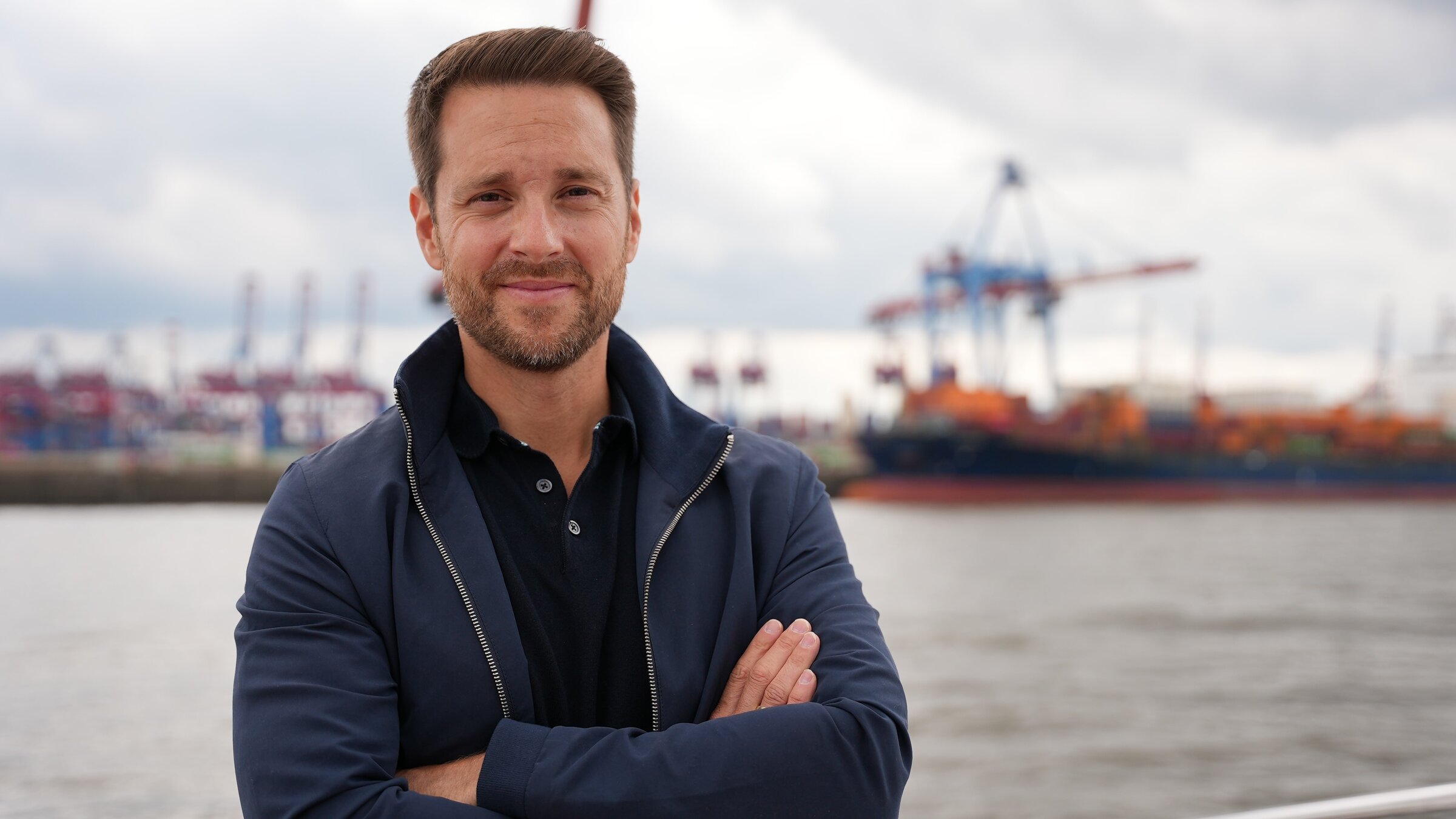 "Terra X: Zeitreise Heimat - Hamburg":  Mirko Drotschmann mit verschränkten Armen, blickt in die Kamera, im Hintergrund Hafenterminal