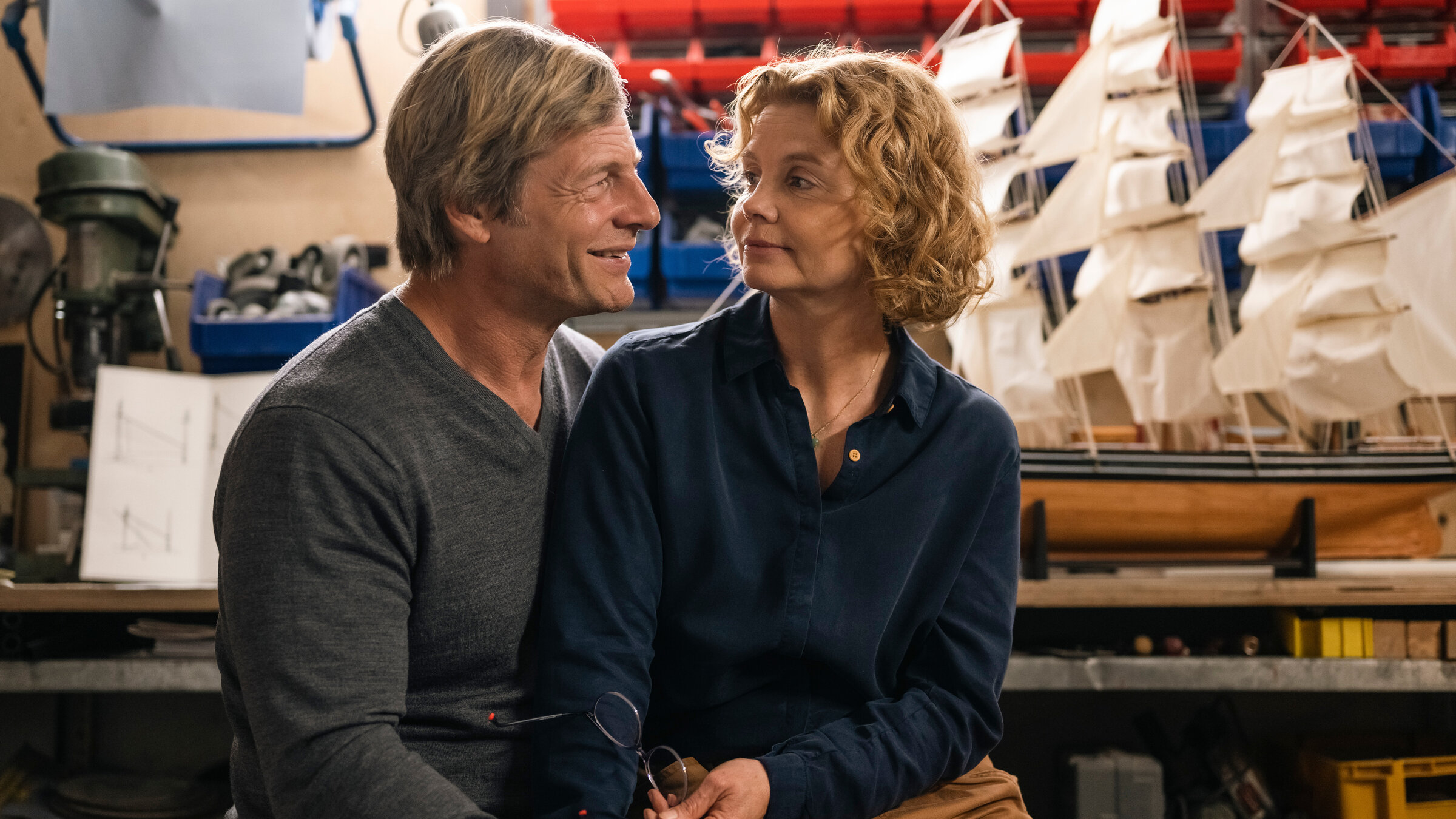 "So weit kommt's noch": In der Werkstatt: Lutz Schmitz (Henning Baum) schaut lächelnd zu seiner Frau Bärbel (Annette Frier), die auf seinem Schoß sitzt und hält sie dabei im Arm. Hinter den beiden steht ein Miniaturschiff.