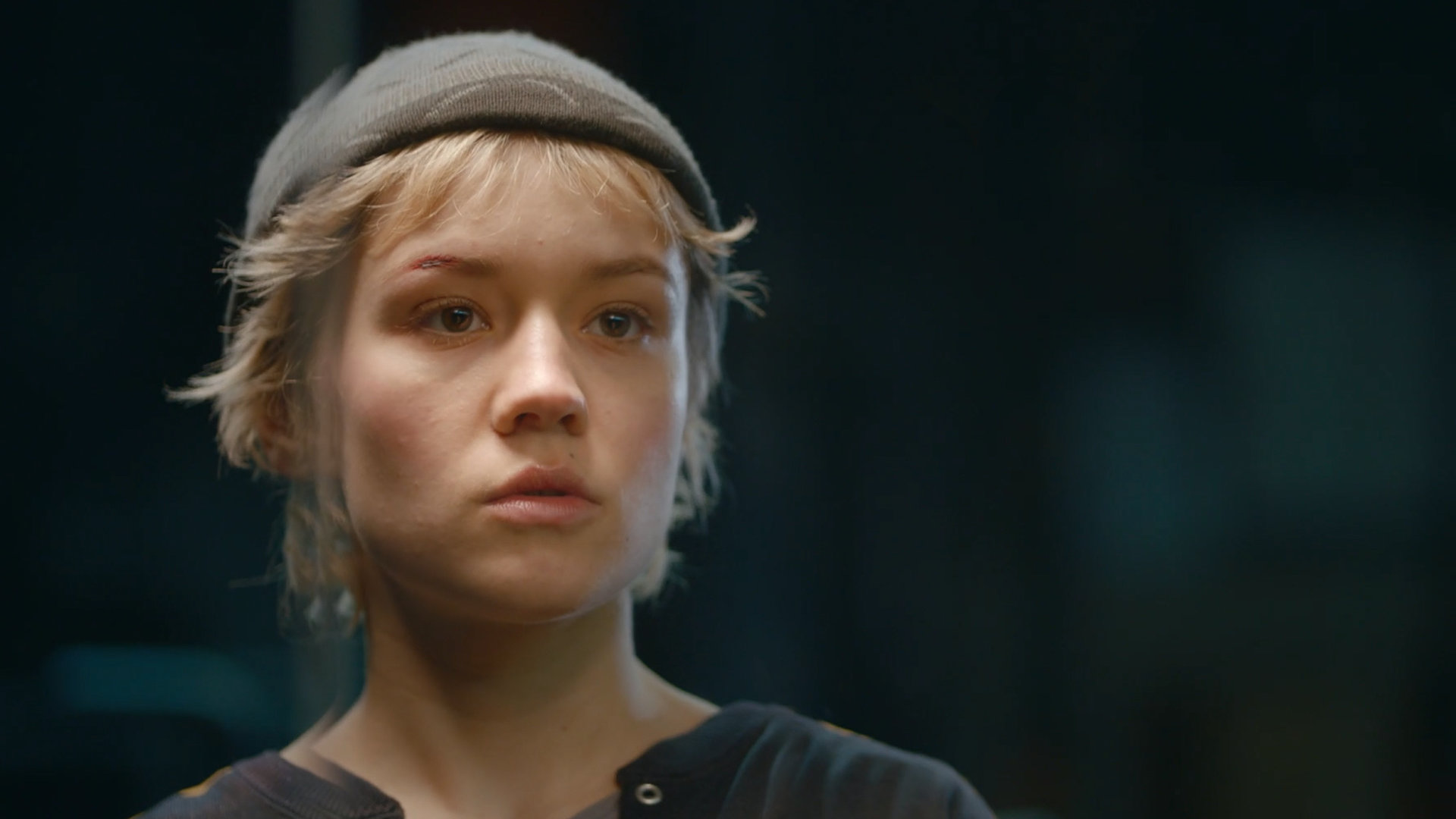 "Becoming Charlie - Mehr als Entweder Oder": Porträt von Charlie (Lea Drinda). Sie trägt eine dunkle Strickmütze und steht vor dunklem Hintergrund.