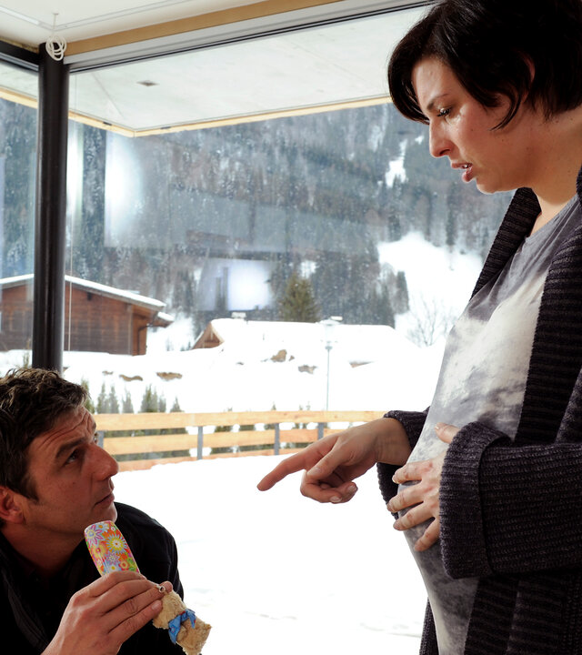 "Der Bergdoktor: Eiszeit": Im Haus der Familie Lechner: Dr. Martin Gruber (Hans Sigl) sitzt in einem Sessel. Während er ein Geschenk in den Händen hält, blickt er zu Maria Lechner (Elena Uhlig) auf. Diese steht, hochschwanger, vor ihm und zeigt mit dem Finger auf das Geschenk. Hinter ihnen gibt eine Fensterfront den Blick auf eine verschneite Landschaft frei.