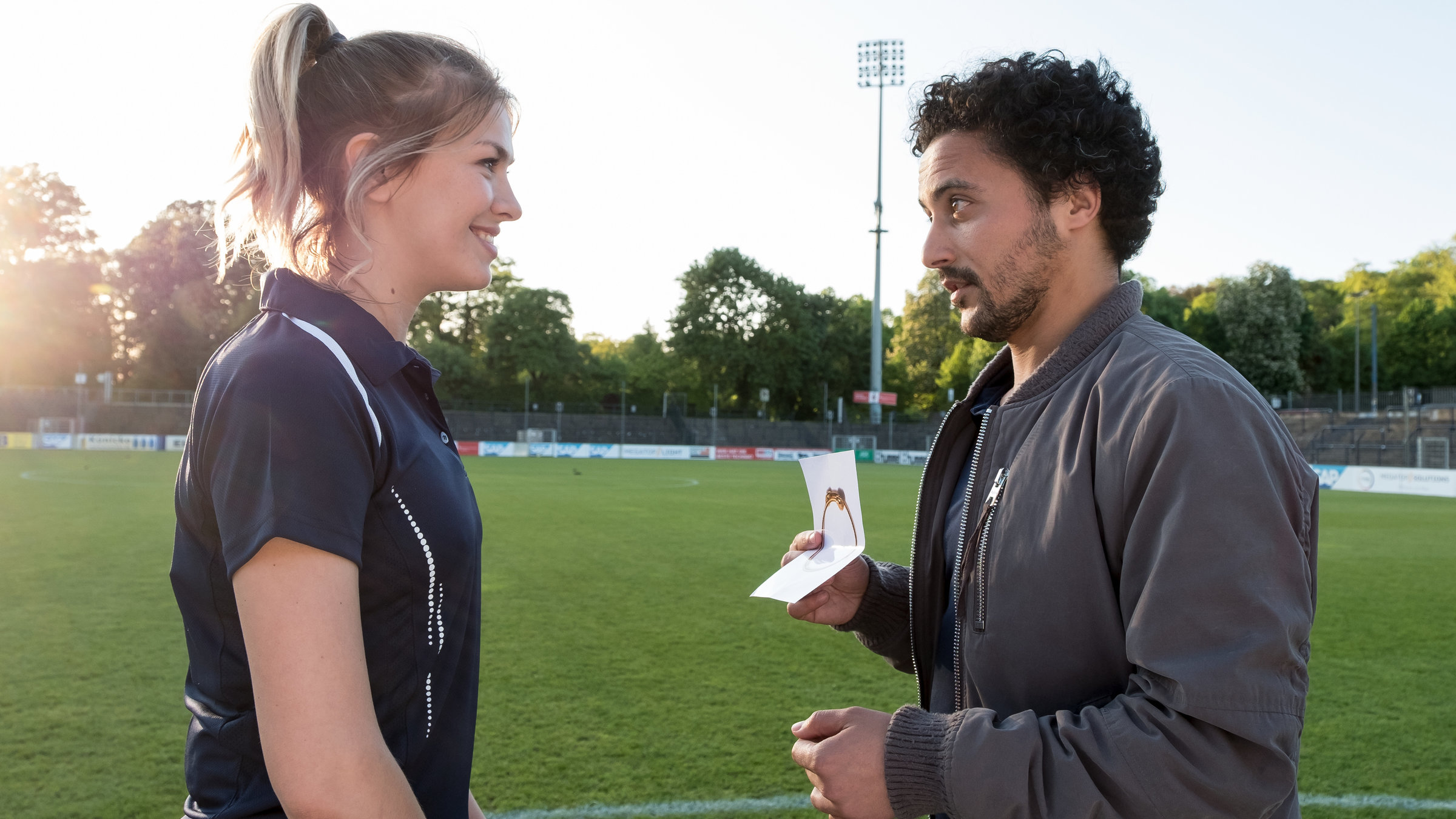 "SOKO Potsdam - Falsches Spiel": Mitten auf dem grünen Fußballfeld von „Potsdam 01“ stehen sich der Kommissar David Grünbaum (Omar El-Saeidi) und die hübsch lächelnde Spielerin Tanja Bell (Zsá Zsá Inci Bürkle), die ihr Vereinstrikot trägt, gegenüber. Grünbaum zeigt ihr ein Foto, auf dem ein Armband abgebildet ist.