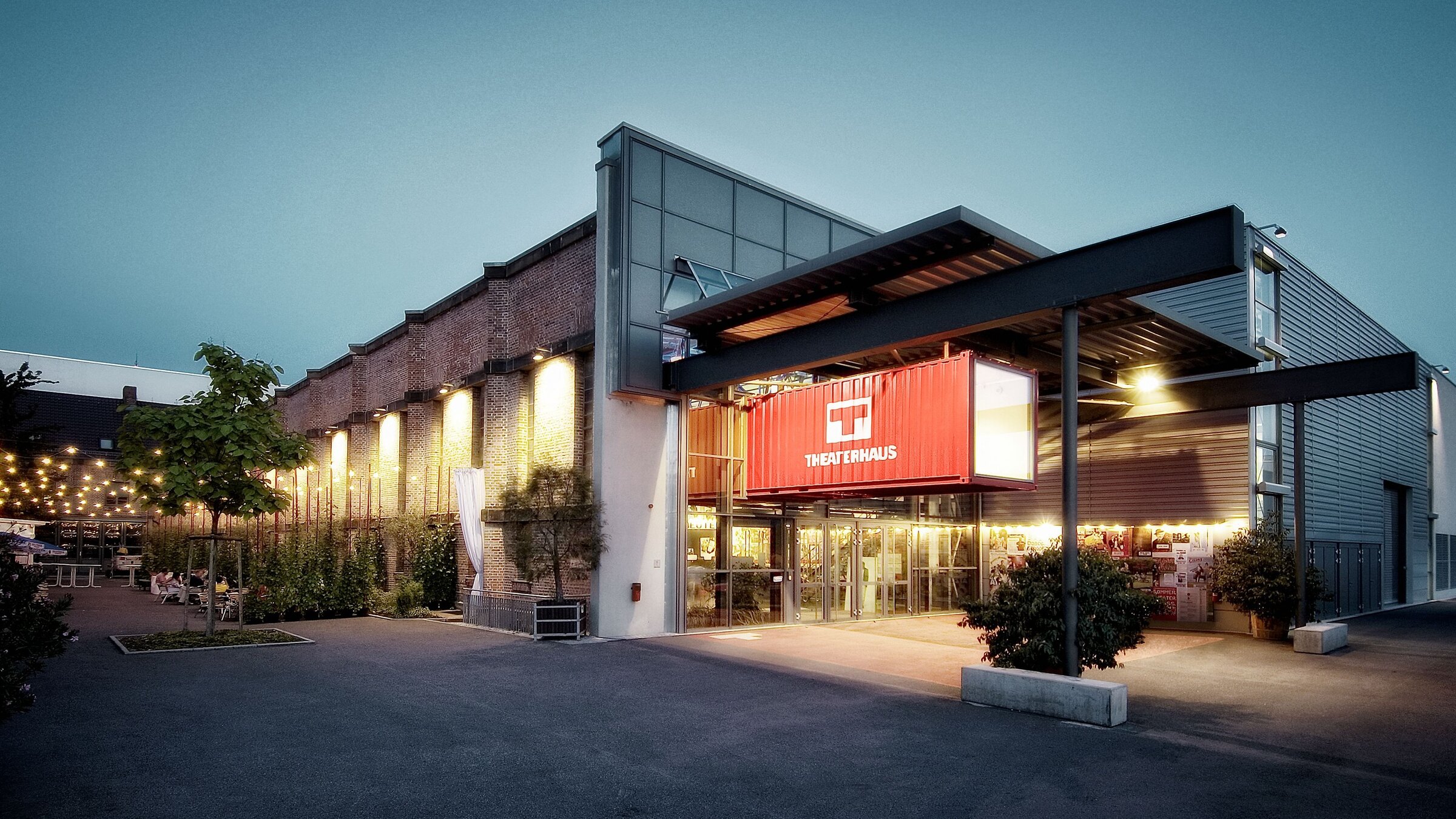 "Der Wert der Kultur – Der Deutsche Theaterpreis DER FAUST": Außenansicht des Theaterhaus Stuttgart in der Dämmerung. Das Gebäude kombiniert Backsteinwände mit moderner Glasarchitektur. Über dem Eingang hängt ein großes rotes Banner mit weißem Logo und Schriftzug „Theaterhaus“. Vor dem Gebäude befindet sich ein breiter, asphaltierter Vorplatz mit Pflanzen in Kübeln. Rechts ragt ein überdachter Eingangsbereich mit Metallträgern hervor, links sind Lichterketten über einer Terrasse sichtbar.