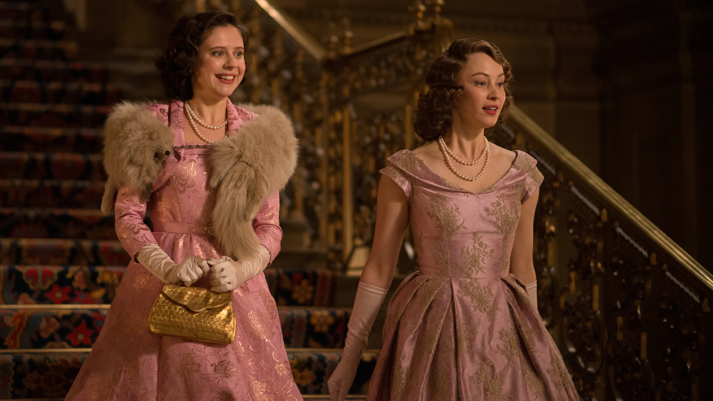 "A Royal Night - Ein königliches Vergnügen": Die beiden Prinzessinnen Margaret (Bel Powley) und Elizabeth (Sarah Gadon) kommen nebeneinander eine prunkvolle Treppe herunter. Sie schauen sich lächelnd um. Beide tragen fast identische, elegante Kleider.